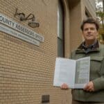 A consulting forester stands beside a New York State government building — a county assessor's office — holding a bound forest management plan document. Professional field attire. The building exterior is clean brick with a small government sign. Autumn trees visible behind. Catskill region, New York. Photorealistic, documentary style, neutral daylight.