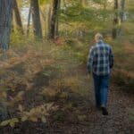 A private landowner in work boots and a flannel shirt walks a trail through an unmanaged northeastern hardwood forest. Japanese barberry shrubs crowd the understory. Dappled morning light filters through an oak and maple canopy. Photorealistic, documentary style, Hudson Valley New York, autumn early light, wide shot.