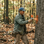 A licensed forester in worn field clothes marking a large oak tree with orange paint in a mixed hardwood forest. Autumn light filtering through the canopy. Other trees visible in background, some marked, some not. Hudson Valley New York forest floor with leaf litter. Documentary photography style, natural light, no filters.