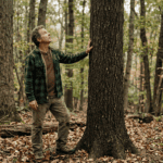 A private landowner in casual outdoor clothes standing in a mature mixed hardwood forest, looking up at a large straight-boled black cherry tree. He has one hand on the bark and appears to be examining the lower stem. Overcast natural light, Hudson Valley New York setting, leaf litter on the ground. Documentary photography style, no filters, warm earth tones. The tree trunk is clearly straight and clean — an obvious high-quality stem.
