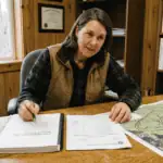 A licensed forester sitting at a wooden desk in a rural office reviewing a forest management plan document and a property invoice side by side. A property map with stand boundaries is spread open nearby. Natural window light, warm interior tones, professional rural setting. Documentary photography style, no filters. The mood is transparent and professional — someone explaining what the work involves and what it costs.