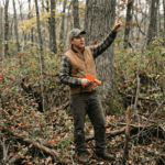 A consulting forester standing in a dense, tangled overgrown woodlot in Sullivan County New York — invasive barberry shrubs visible in the understory, competing small-diameter stems crowding a larger oak tree behind him. He holds a marking paint gun and points toward the oak crown, identifying it as a crop tree. Overcast natural forest light, muted earth tones. Documentary photography style, no filters. The mood is purposeful — someone reading chaos and finding the recoverable structure within it.