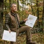 A private landowner in casual outdoor clothes standing in a mature mixed hardwood forest in Sullivan County New York, holding an open property tax bill in one hand and a forest management plan document in the other. His expression is calm and informed — someone who understands what the document in his hand is saving him. Overcast natural forest light, warm earth tones, no filters. Documentary photography style. Stone wall visible at the forest edge in the background.