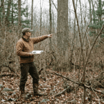 A forester in field clothes standing inside a dense, crowded hardwood woodlot in upstate New York. The understory is thick with competing shrubs and small-diameter stems. He is pointing toward a tall, well-formed oak tree partially hidden by surrounding growth. Overcast forest light, late autumn, leaves on the ground. Documentary photography style, natural color, no filters.