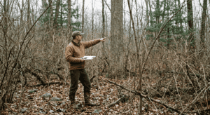 A forester in field clothes standing inside a dense, crowded hardwood woodlot in upstate New York. The understory is thick with competing shrubs and small-diameter stems. He is pointing toward a tall, well-formed oak tree partially hidden by surrounding growth. Overcast forest light, late autumn, leaves on the ground. Documentary photography style, natural color, no filters.