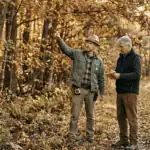 A licensed forester and a private landowner standing together at the edge of a mature mixed hardwood forest in Sullivan County New York. The forester points toward the canopy while the landowner listens and takes notes on a small pad. Morning light, professional but approachable mood — a first property walk in progress. Documentary photography style, natural color, warm earth tones, no filters. Neither figure is staged — both look like working professionals engaged in a real conversation.
