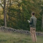 A first-time landowner — dressed in casual outdoor clothes, clearly not a professional forester — standing at the edge of a mature mixed hardwood forest in upstate New York, looking into the woods with a small notebook in hand. Morning light, slightly uncertain posture — someone seeing their woodland for the first time with fresh eyes. Documentary photography style, warm earth tones, no filters. Stone wall visible at the forest edge suggesting old farmland.