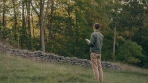 A first-time landowner — dressed in casual outdoor clothes, clearly not a professional forester — standing at the edge of a mature mixed hardwood forest in upstate New York, looking into the woods with a small notebook in hand. Morning light, slightly uncertain posture — someone seeing their woodland for the first time with fresh eyes. Documentary photography style, warm earth tones, no filters. Stone wall visible at the forest edge suggesting old farmland.