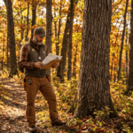 Licensed forester in field gear reviewing a paper document in a mature Hudson Valley hardwood forest, autumn foliage, tall oak and maple trees, dappled golden afternoon light filtering through orange and yellow leaves, realistic professional photography style, no text overlay, New York countryside setting