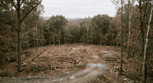 A realistic, editorial-style outdoor photograph on a rural wooded property in the Hudson Valley region of New York, taken in soft overcast daylight. The scene shows a partially cleared building site: a rectangular section of forest has been opened up with visible stumps and cut logs stacked to one side, marking out a building envelope, while dense standing hardwood forest remains at the edges of the frame. Survey stakes with orange flagging tape mark the corners of the cleared area. A small access path or rough driveway cut leads into the site from the foreground. Muted natural color palette — browns, dull greens, gray sky, no oversaturation. Shot from a slightly elevated three-quarter angle to show the full clearing footprint against the remaining tree line. Photorealistic, documentary/photojournalistic style, natural lens compression. No people in frame. No text, no logos, no watermarks, no signage.